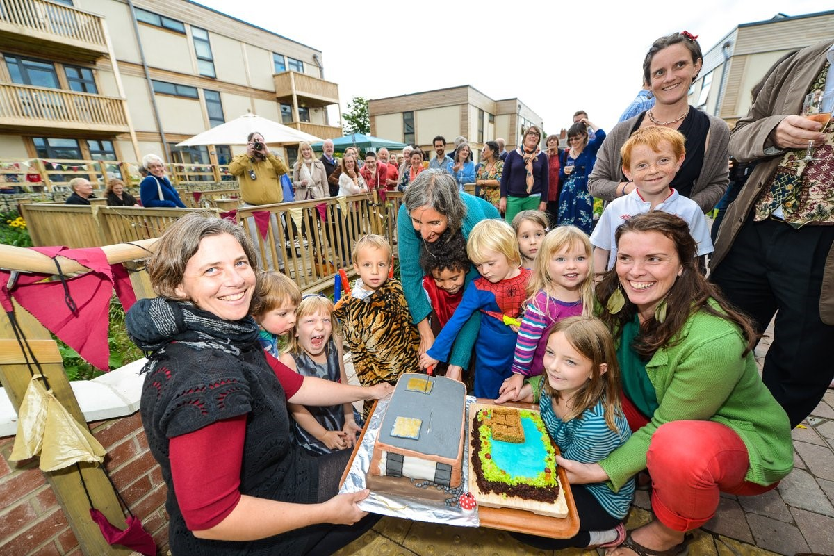 An outdoor birthday party, with a group holding a cake in the foreground, lots of onlookers behind.