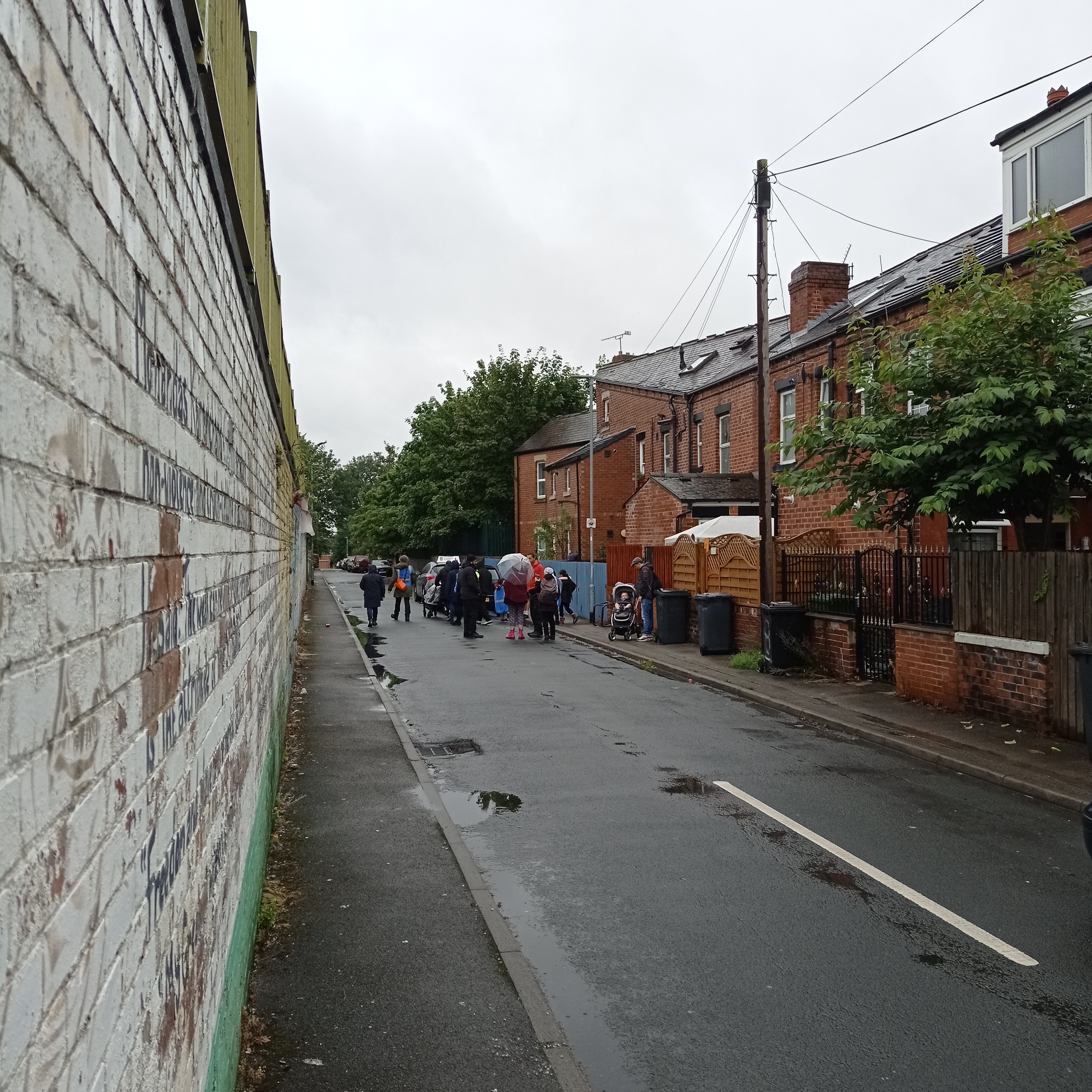 A cluster of people stand at the far end of a long straight back street with the backs of red brick terraced houses on one side and a white-painted brick wall on the other.